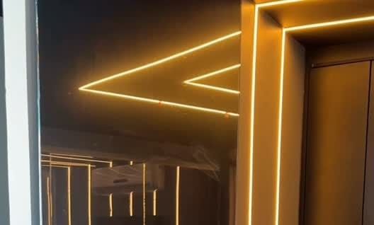 Porta de vidro escuro do showroom, com linhas de LED no teto refletidas e uma moldura de luz quente em volta da passagem